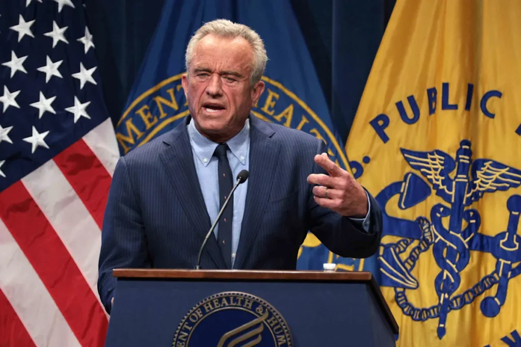 RFK Jr. speaks at a podium with microphones, standing in front of U.S. and Department of Health and Human Services flags.
