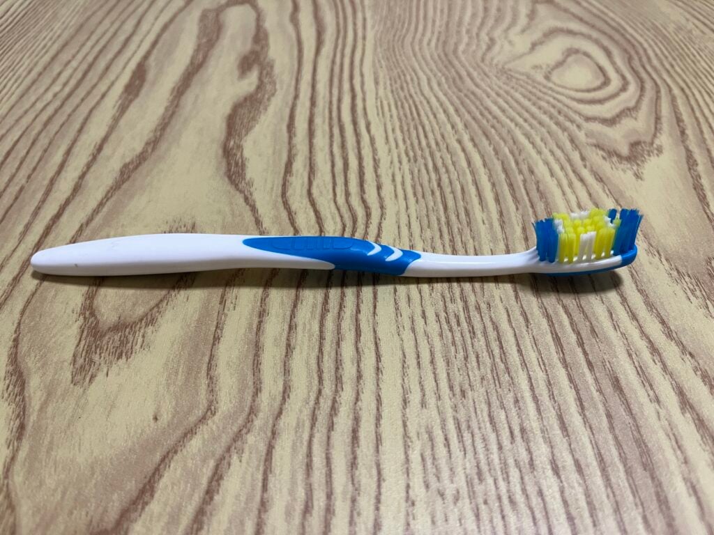 A white and blue toothbrush with yellow and blue bristles lies on a wood-patterned surface.