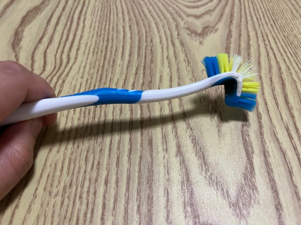 A person holds a bent cleaning brush with blue, yellow, and white bristles on a light wood-grain surface.