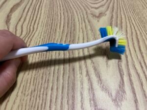 A person holds a bent cleaning brush with blue, yellow, and white bristles on a light wood-grain surface.