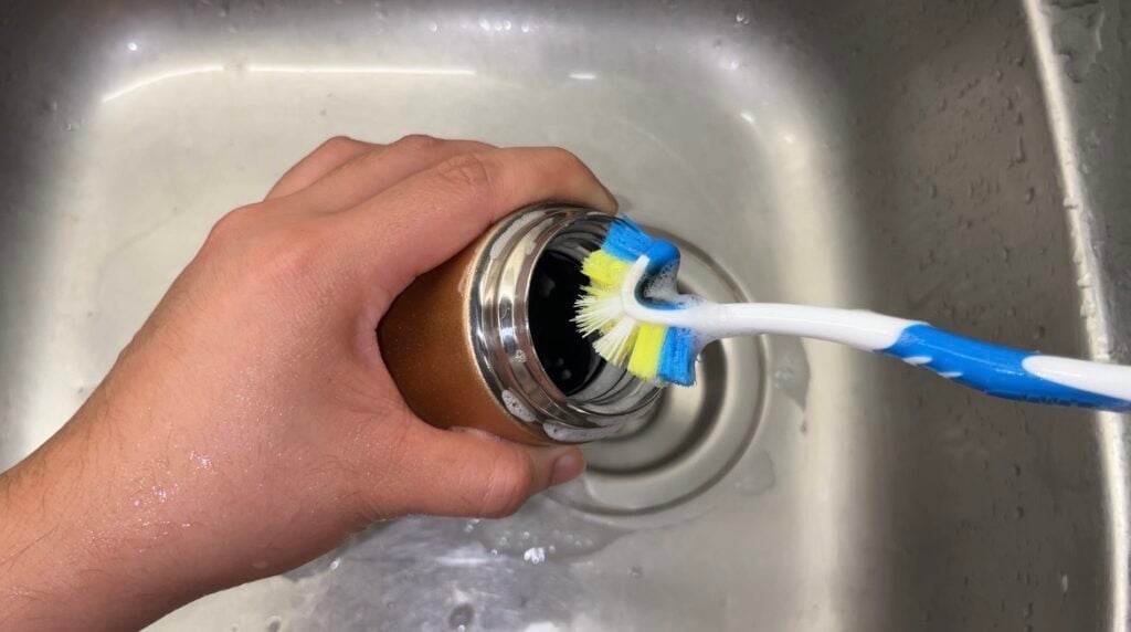 A person uses a toothbrush to clean the inside rim of a metal tumbler over a kitchen sink.