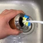A person uses a toothbrush to clean the inside rim of a metal tumbler over a kitchen sink.