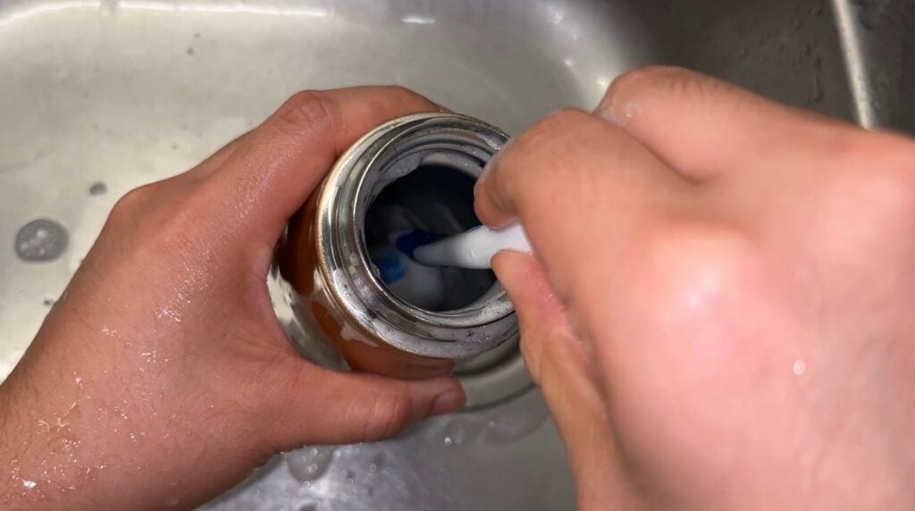 A person uses a brush to clean the inside of a metal bottle over a sink.