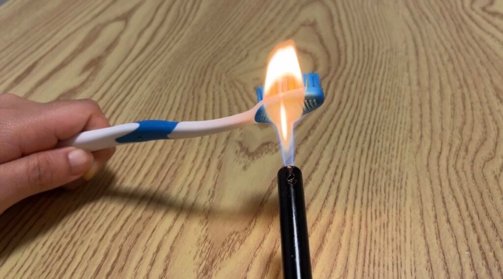 A hand holds a toothbrush over a lighter, with the toothbrush head being exposed to an open flame above a wood-patterned surface.