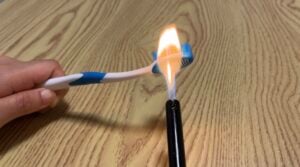 A hand holds a toothbrush over a lighter, with the toothbrush head being exposed to an open flame above a wood-patterned surface.
