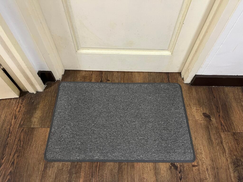 A gray rectangular doormat lies on a wooden floor in front of a closed white door.
