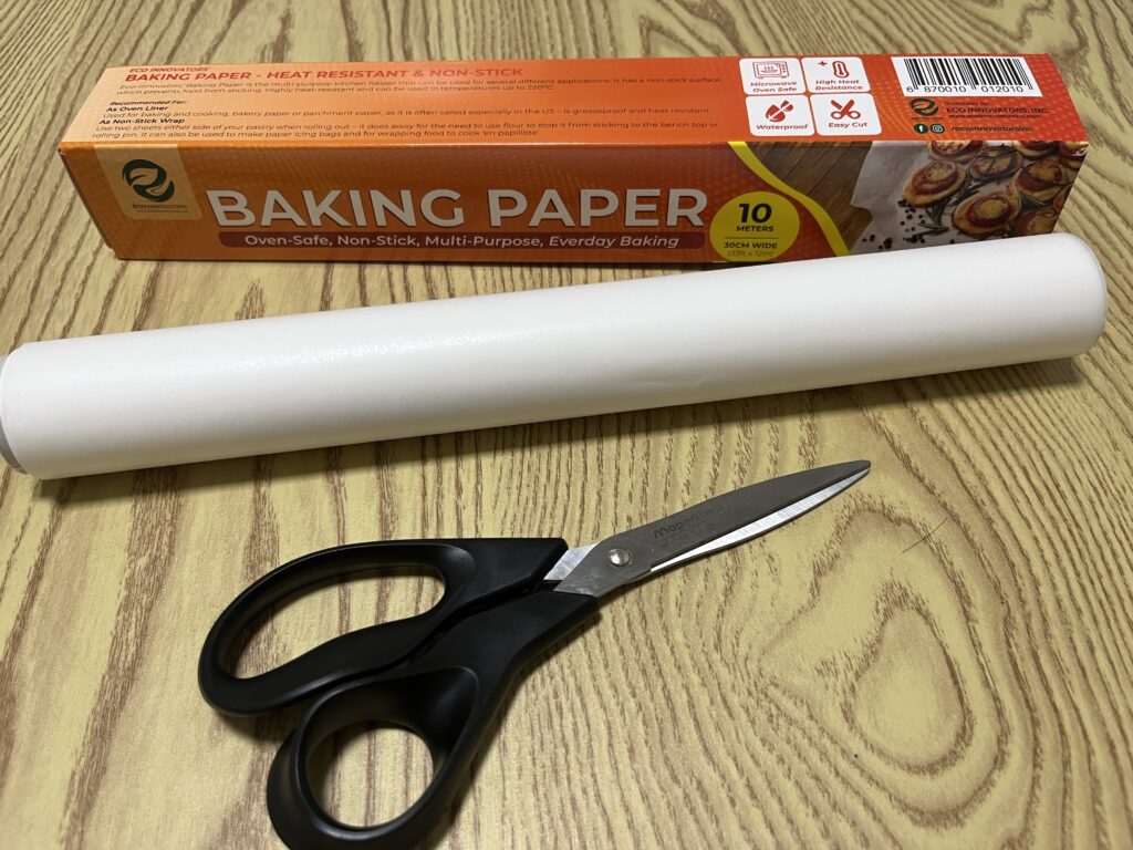 A box of baking paper, a roll of baking paper, and a pair of black scissors on a wooden surface.