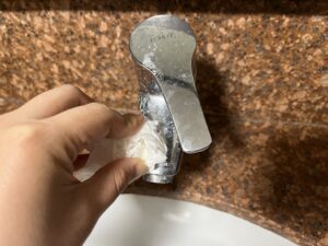 A hand uses a tissue to clean a silver sink faucet mounted on a brown speckled countertop.