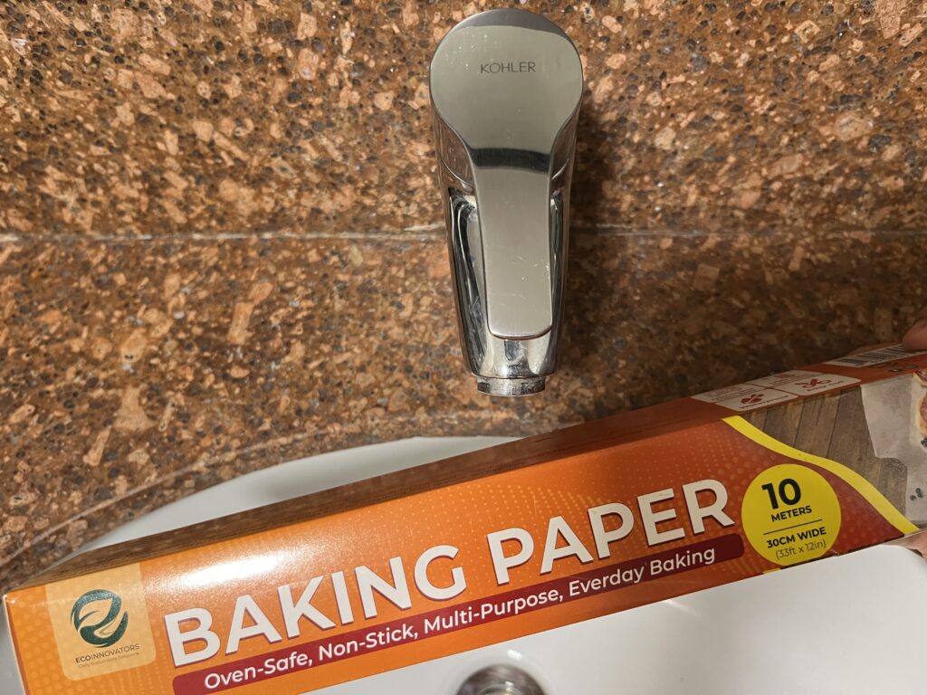 A box of baking paper held over a sink with a silver faucet and brown tiled background.