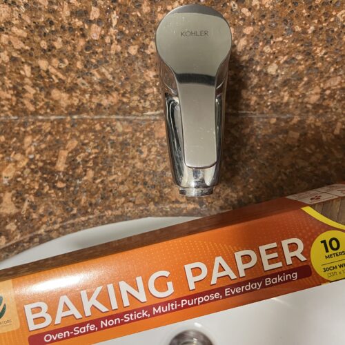 A box of baking paper held over a sink with a silver faucet and brown tiled background.