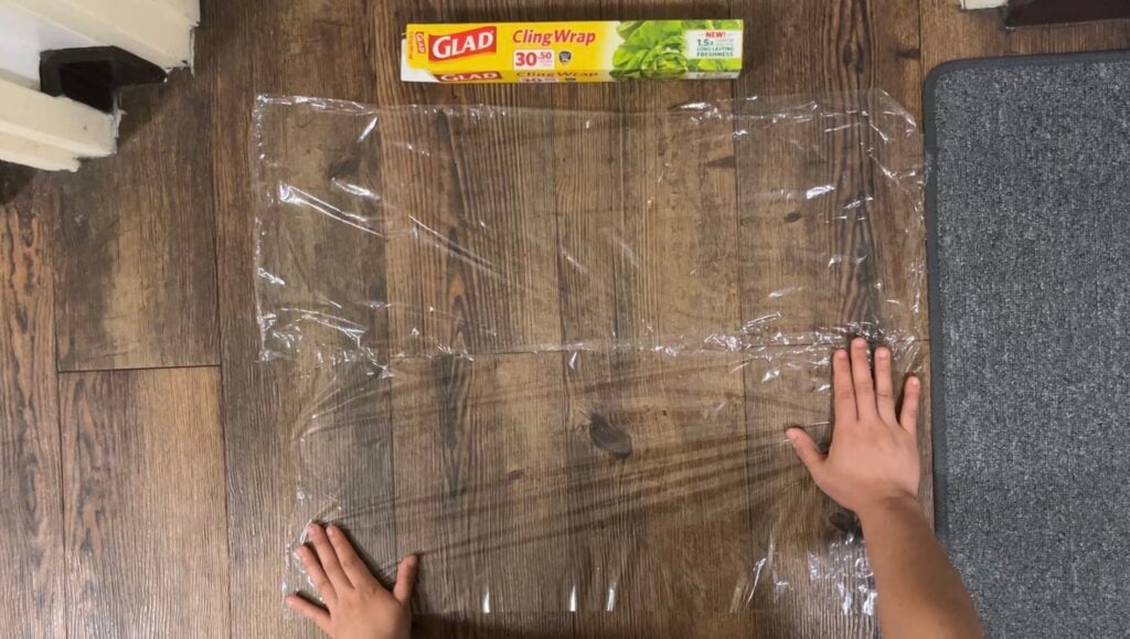 A person unrolling a sheet of Glad Cling Wrap on a wooden surface, with the packaging box placed above the plastic wrap.