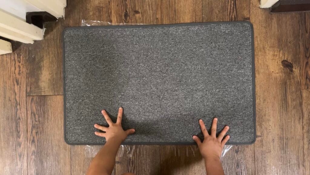 A pair of small hands pressing down on a rectangular gray mat placed on a wooden floor.