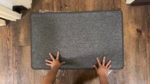 A pair of small hands pressing down on a rectangular gray mat placed on a wooden floor.