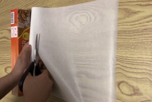 A person uses scissors to cut a sheet of white parchment paper on a wooden surface, with a box of parchment paper visible in the background.