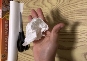 A hand holding a crumpled piece of white paper, with scissors, baking paper, and a napkin on a wooden surface in the background.