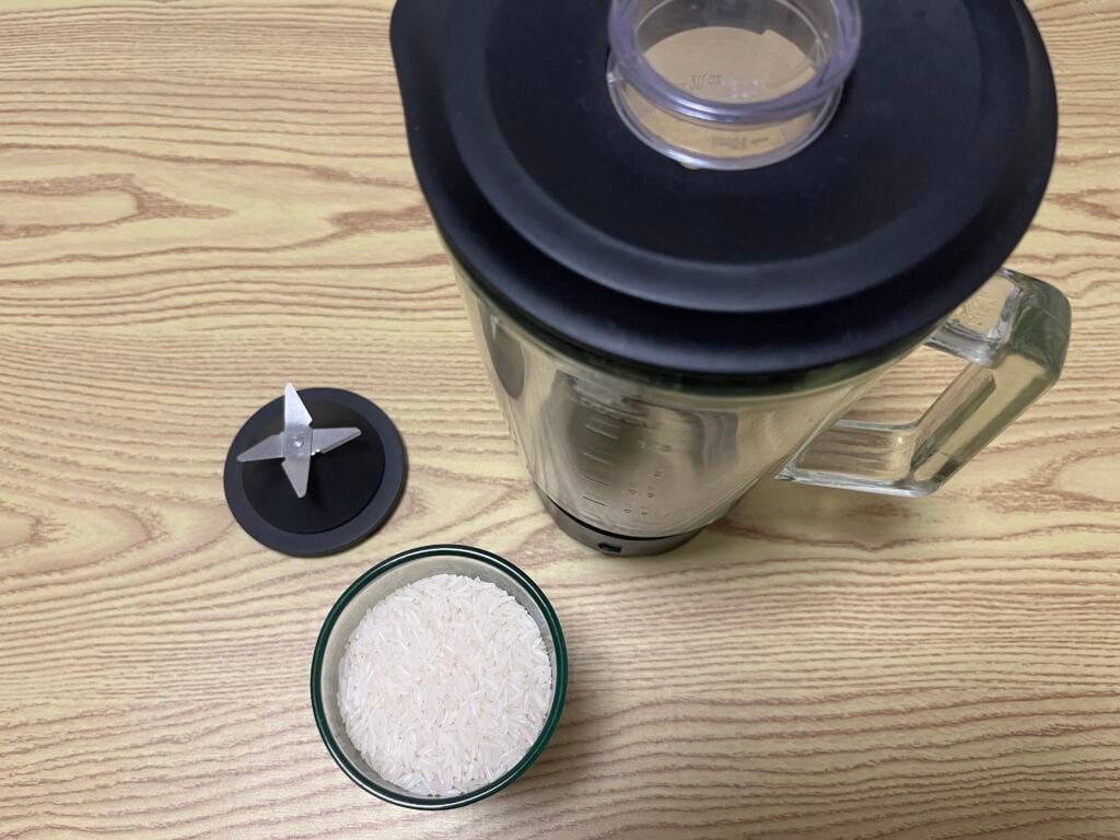 A blender with its lid removed, a detached metal blade, and a bowl of uncooked white rice are placed on a wooden surface.