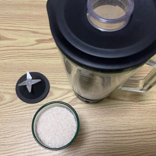 A blender with its lid removed, a detached metal blade, and a bowl of uncooked white rice are placed on a wooden surface.