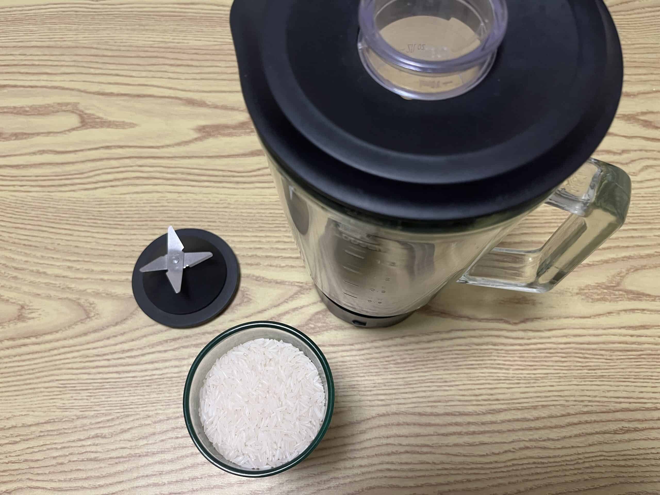 A blender with its lid removed, a detached metal blade, and a bowl of uncooked white rice are placed on a wooden surface.