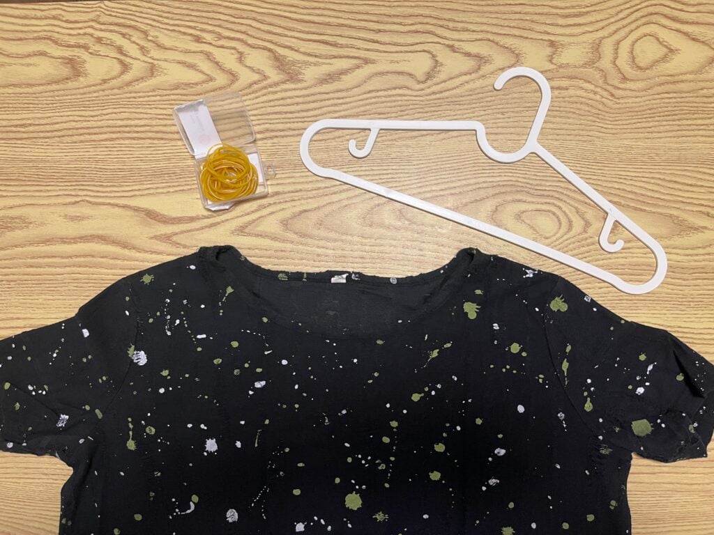 A black shirt with white and green splatters, a clear tape dispenser with yellow tape, and a white plastic hanger are on a light wood surface.