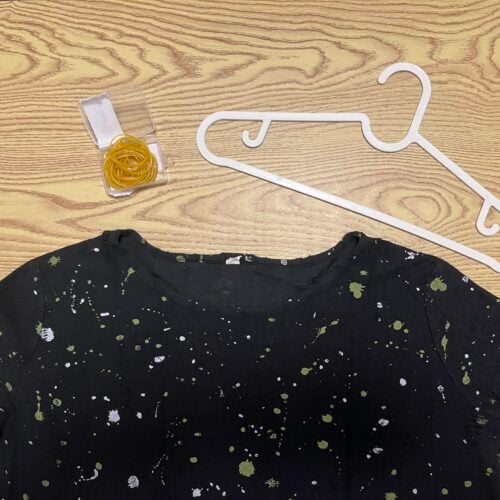 A black shirt with white and green splatters, a clear tape dispenser with yellow tape, and a white plastic hanger are on a light wood surface.