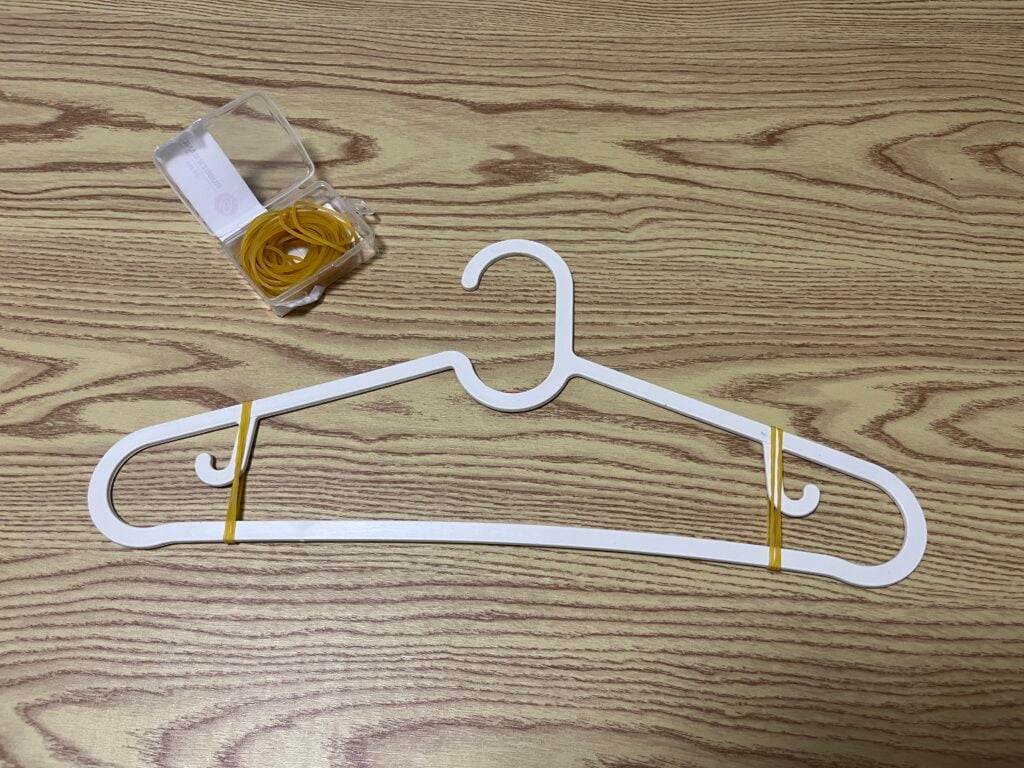 A white plastic clothes hanger with two rubber bands wrapped around it lies on a wood-patterned surface. A small plastic box containing more rubber bands is nearby.