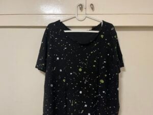 A black T-shirt with green and white paint splatter patterns hangs on a white plastic hanger against a cream-colored closet.