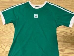 Green Adidas T-shirt with white collar, sleeve trim, and shoulder stripes, displayed on a light wood surface. Adidas logo patch centered on the chest.