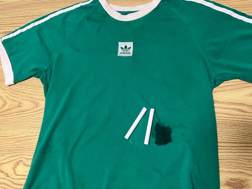 A green Adidas t-shirt with a dark stain and three pieces of white chalk lying on it, placed on a wooden surface.