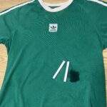 A green Adidas t-shirt with a dark stain and three pieces of white chalk lying on it, placed on a wooden surface.