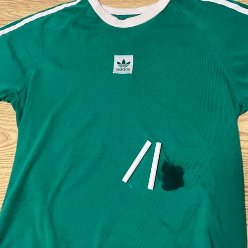 A green Adidas t-shirt with a dark stain and three pieces of white chalk lying on it, placed on a wooden surface.