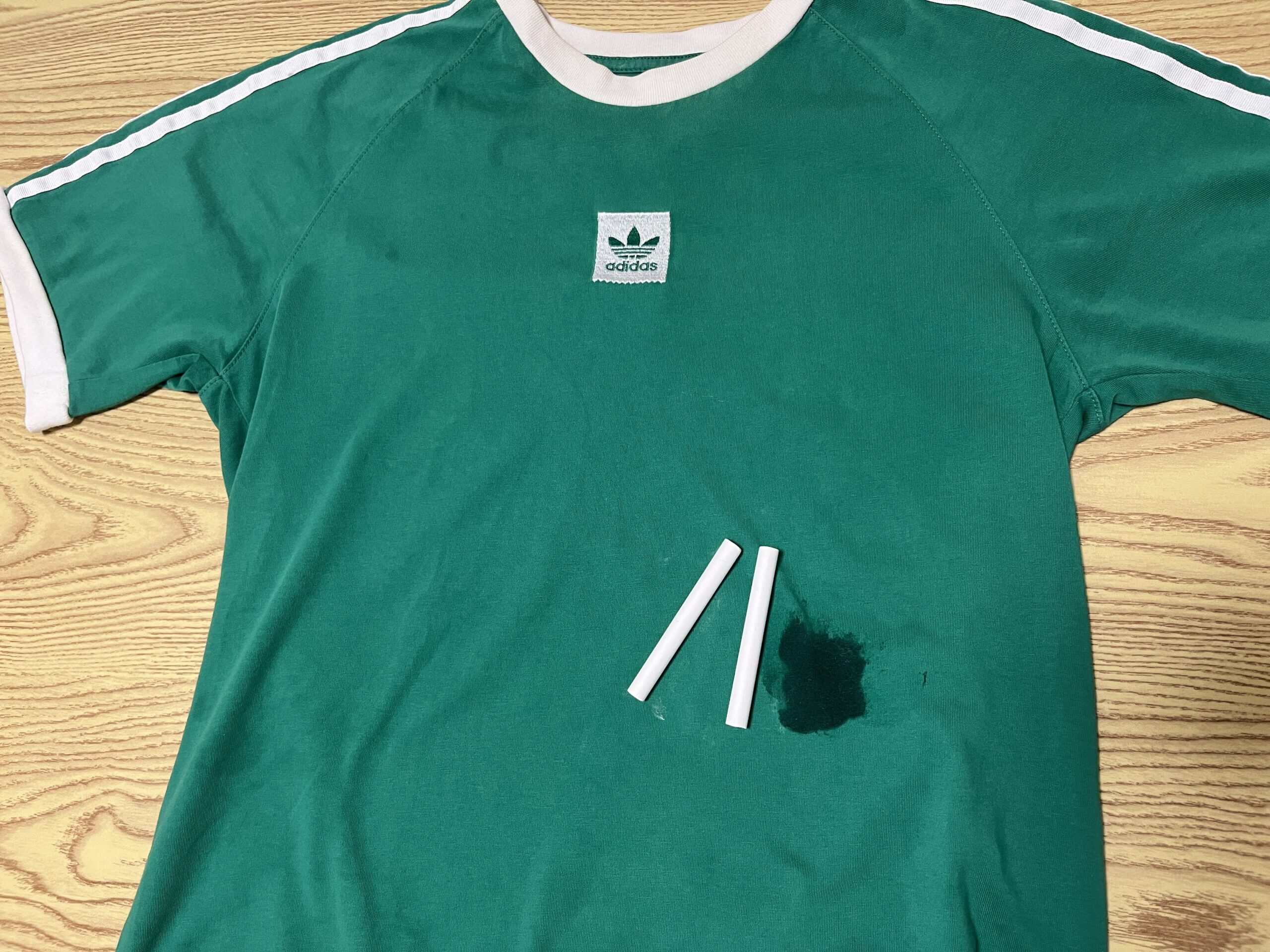 A green Adidas t-shirt with a dark stain and three pieces of white chalk lying on it, placed on a wooden surface.