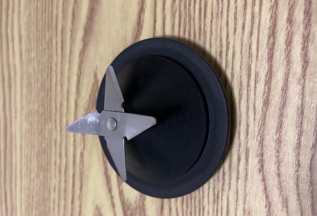 A metal blender blade assembly with two sharp blades is attached to a black plastic base, placed on a wooden surface.