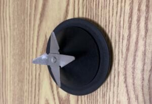 A metal blender blade assembly with two sharp blades is attached to a black plastic base, placed on a wooden surface.