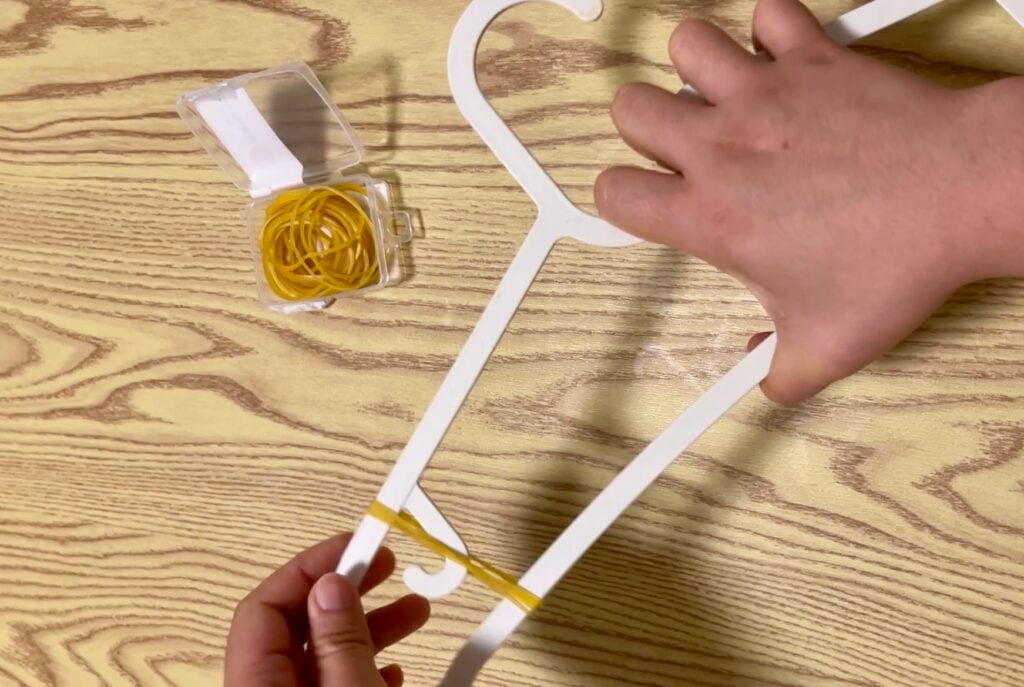 Two hands stretch a yellow rubber band around the bottom of a white clothes hanger on a wood-patterned surface. An open box of yellow rubber bands is nearby.