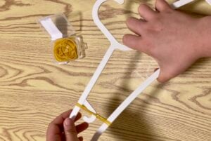Two hands stretch a yellow rubber band around the bottom of a white clothes hanger on a wood-patterned surface. An open box of yellow rubber bands is nearby.