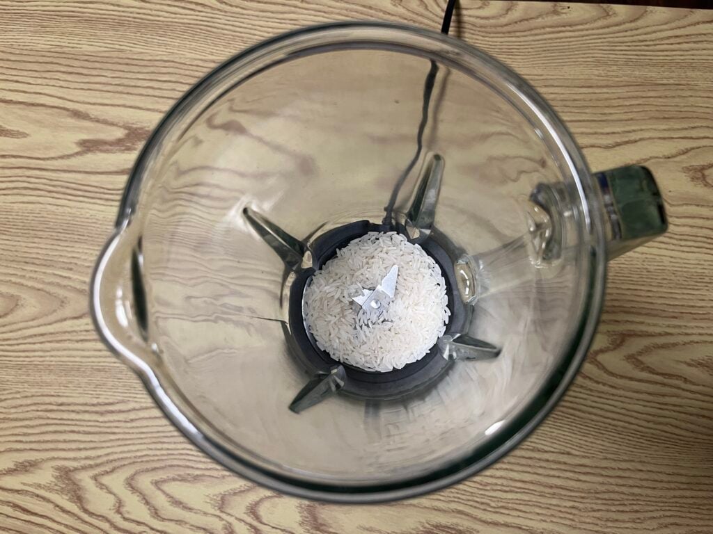 Overhead view of uncooked white rice inside a glass blender jar on a light wood surface.