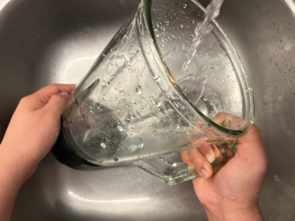 A person rinses a glass blender pitcher under running water in a stainless steel sink.