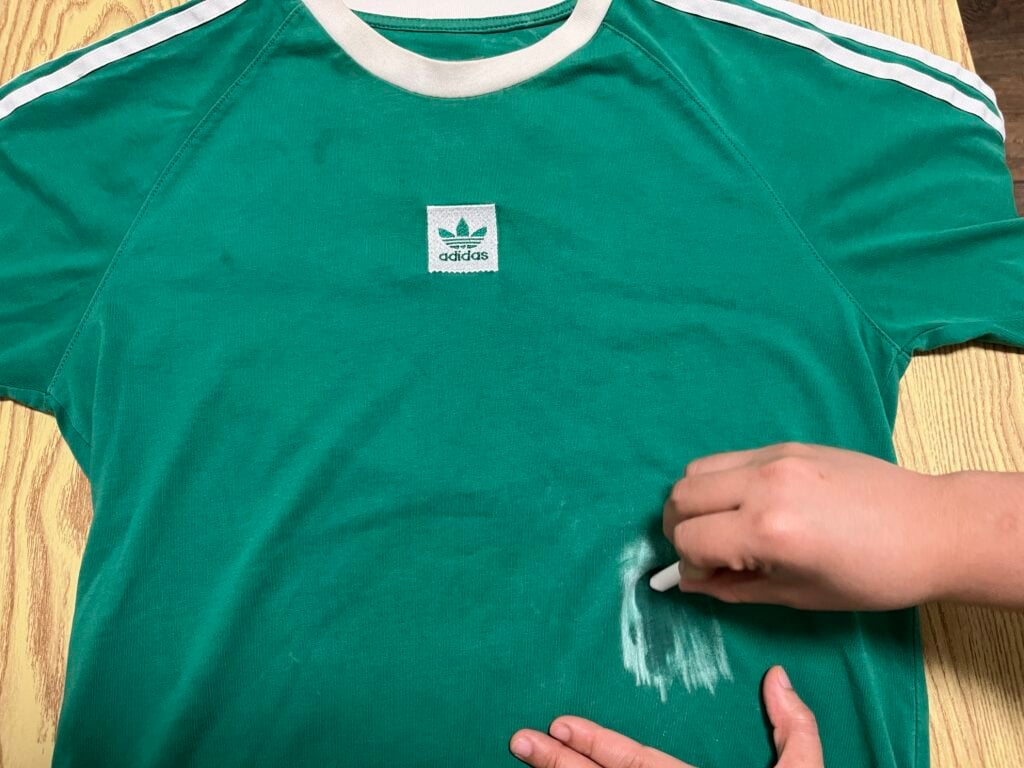 A person rubs a white substance onto a green Adidas t-shirt, appearing to clean or treat a stain on the fabric.