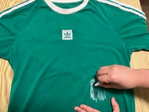 A person rubs a white substance onto a green Adidas t-shirt, appearing to clean or treat a stain on the fabric.