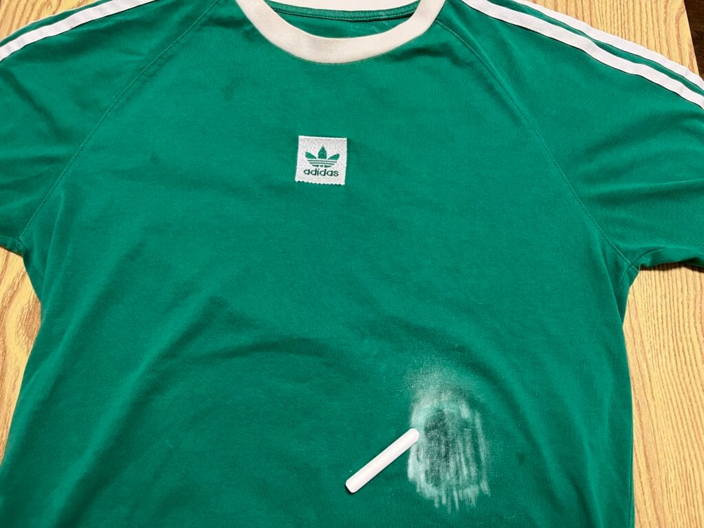 A green Adidas t-shirt with a white stripe, lying on a wooden surface, has a white chalk stick and powdery chalk smudge on the lower right side.