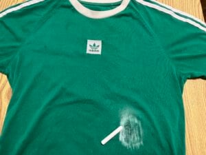 A green Adidas t-shirt with a white stripe, lying on a wooden surface, has a white chalk stick and powdery chalk smudge on the lower right side.
