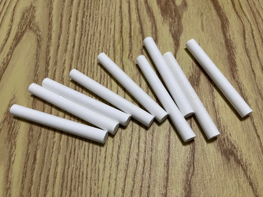 Ten white chalk sticks are scattered on a light brown wooden surface with a visible grain pattern.