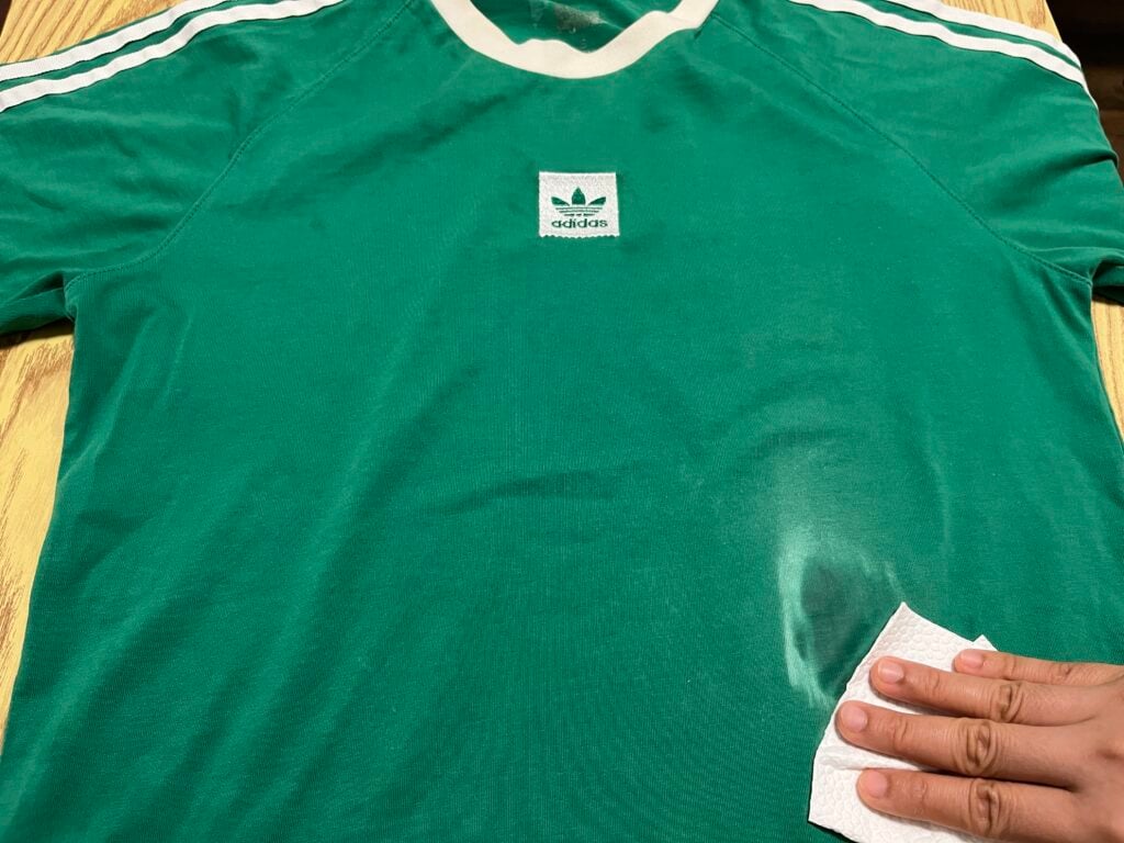 A hand wipes the right side of a green Adidas shirt with white stripes and collar, which is laid flat on a wooden surface.