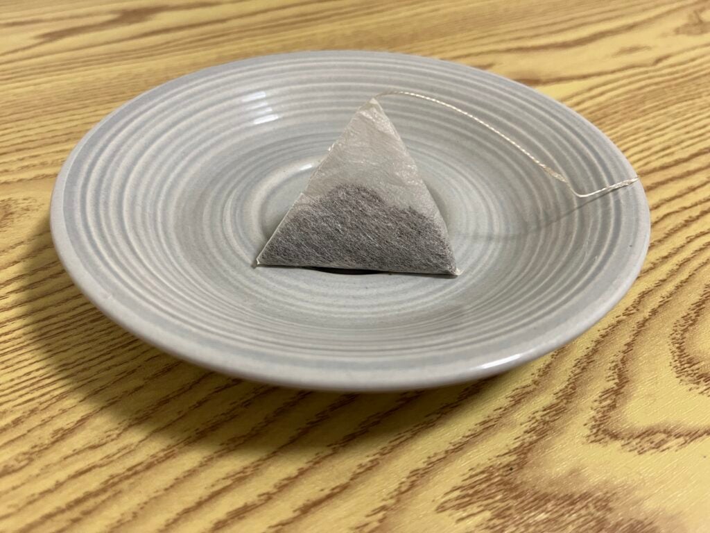 A pyramid-shaped tea bag sits on a round, light gray saucer atop a wooden surface.