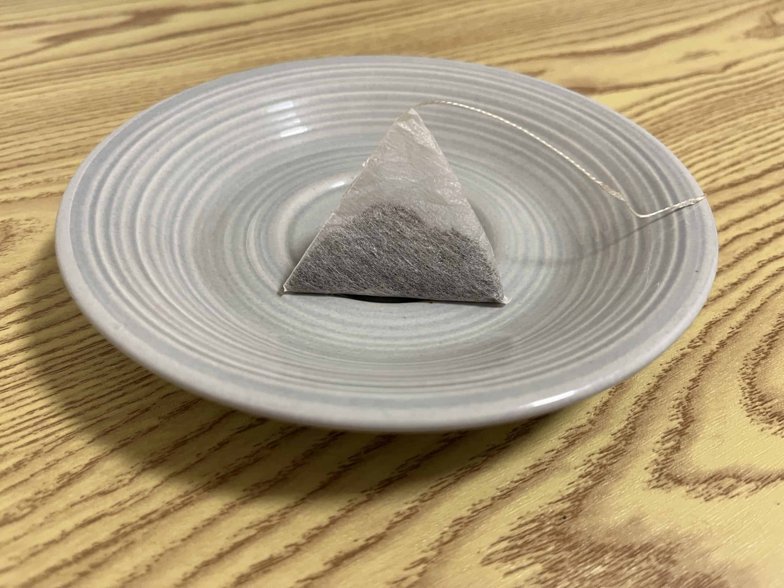 A pyramid-shaped tea bag sits on a round, light gray saucer atop a wooden surface.