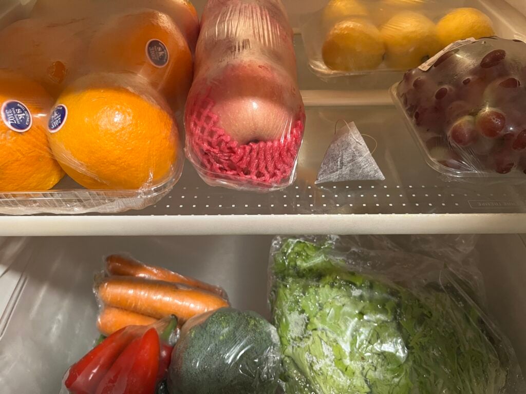 Refrigerator shelves with oranges, apples, lemons, grapes, carrots, lettuce, bell peppers, and a broccoli, all wrapped in plastic.