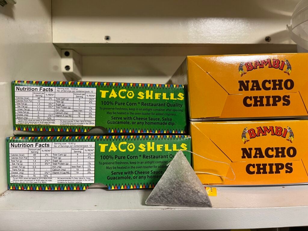 Boxes of taco shells and two boxes of nacho chips are displayed on a white shelf, with a triangular metal shelf divider in front of the taco shell boxes.