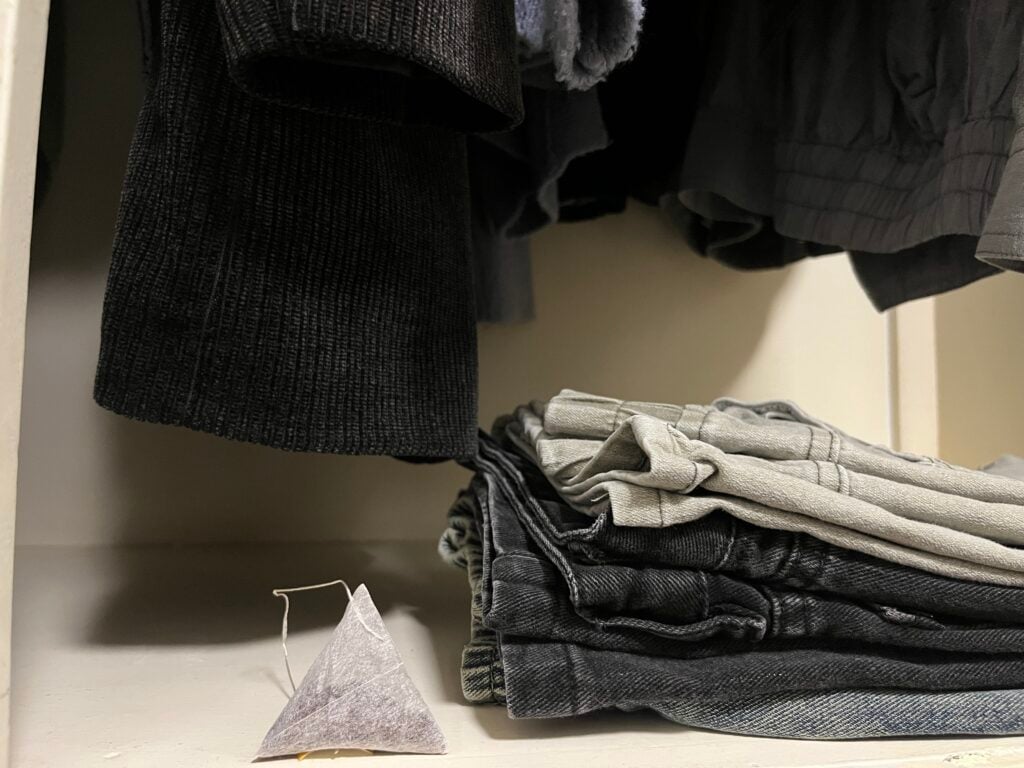 A small pyramid-shaped sachet sits next to a folded stack of gray and black jeans inside a closet with dark clothing hanging above.