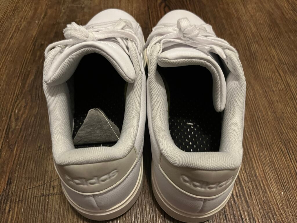 A pair of white Adidas sneakers with laces viewed from above, placed side by side on a wooden floor.