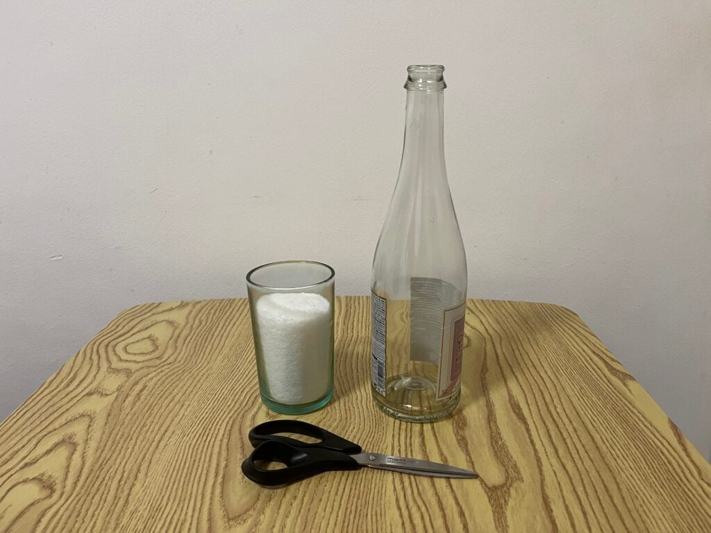 A pair of black scissors, an empty glass bottle, and a glass filled with white granules sit on a wooden table against a plain wall.